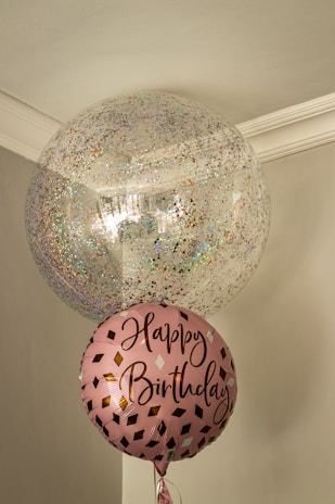 Two balloons are floating indoors, one large and transparent with multicolored confetti inside, and one smaller pink balloon with the words 'Happy Birthday' written on it in a stylish script. The setting is a corner of a room with muted colors and a simple ceiling molding.