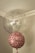 Two balloons are floating indoors, one large and transparent with multicolored confetti inside, and one smaller pink balloon with the words 'Happy Birthday' written on it in a stylish script. The setting is a corner of a room with muted colors and a simple ceiling molding.