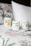 Three candles in minimalist white containers are placed on a bed with a floral-patterned quilt and a plain white pillow. The candle labels display the brand 'Clare Makes' along with colorful abstract designs and text describing scents.