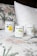 Three candles in minimalist white containers are placed on a bed with a floral-patterned quilt and a plain white pillow. The candle labels display the brand 'Clare Makes' along with colorful abstract designs and text describing scents.