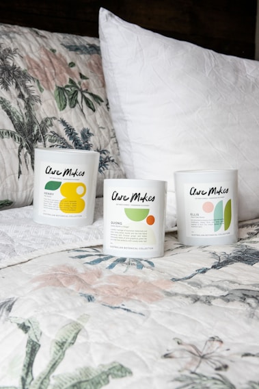 Three candles in minimalist white containers are placed on a bed with a floral-patterned quilt and a plain white pillow. The candle labels display the brand 'Clare Makes' along with colorful abstract designs and text describing scents.