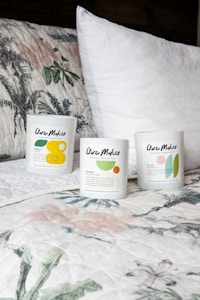 Three candles in minimalist white containers are placed on a bed with a floral-patterned quilt and a plain white pillow. The candle labels display the brand 'Clare Makes' along with colorful abstract designs and text describing scents.