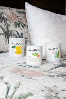 Three candles in minimalist white containers are placed on a bed with a floral-patterned quilt and a plain white pillow. The candle labels display the brand 'Clare Makes' along with colorful abstract designs and text describing scents.