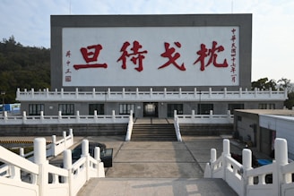 A large structure featuring a massive billboard with bold red Chinese characters displayed prominently against a gray background. The building has an architectural design that includes multiple white railings and stairs leading up to it. Trees and greenery surround the area, indicating a possibly rural or suburban setting.
