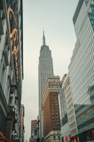A tall skyscraper rises prominently among other high-rise buildings in an urban setting. The architecture features large windows and classic design elements, with other modern buildings lining the street.