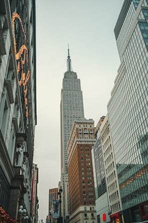 A tall skyscraper rises prominently among other high-rise buildings in an urban setting. The architecture features large windows and classic design elements, with other modern buildings lining the street.
