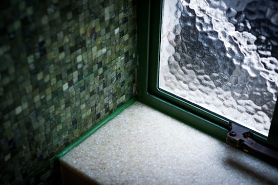 A section of a room interior with a small window. The window has frosted glass and a green frame. The walls are covered with small, green-toned mosaic tiles. A smooth beige surface, possibly a countertop or windowsill, is visible beneath the window.