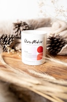 A white candle labeled 'Clare Makes' sits on a wooden surface, surrounded by dried pine cones and beige textured fabric. Delicate branches with small white flowers and wisps of dried grass add an earthy touch, creating a cozy, natural atmosphere.