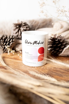 A white candle labeled 'Clare Makes' sits on a wooden surface, surrounded by dried pine cones and beige textured fabric. Delicate branches with small white flowers and wisps of dried grass add an earthy touch, creating a cozy, natural atmosphere.