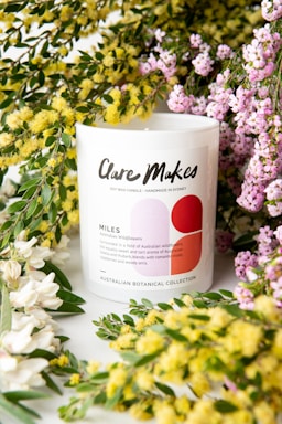 A white candle jar with a label reading 'Clare Makes' and 'Miles' is surrounded by vibrant yellow and purple flowers. The label mentions the scent as reminiscent of Australian wildflowers and has a graphic design with red and pink circles. Green leaves enhance the floral arrangement, creating a fresh and natural setting.