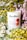 A white candle jar with a label reading 'Clare Makes' and 'Miles' is surrounded by vibrant yellow and purple flowers. The label mentions the scent as reminiscent of Australian wildflowers and has a graphic design with red and pink circles. Green leaves enhance the floral arrangement, creating a fresh and natural setting.
