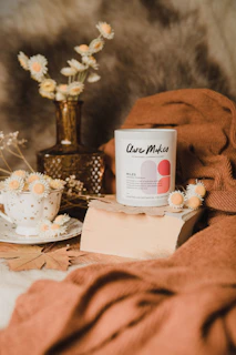 A candle with the label 'Clare Makes' is placed on a piece of wood. Surrounding it are dried flowers in a white polka dot cup and a brown glass vase. A soft brown fabric drapes around the setting, creating a cozy, natural aesthetic.