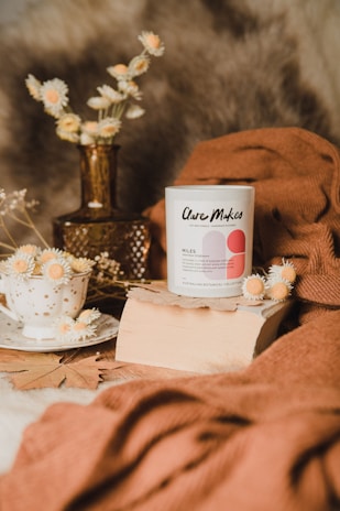 A candle with the label 'Clare Makes' is placed on a piece of wood. Surrounding it are dried flowers in a white polka dot cup and a brown glass vase. A soft brown fabric drapes around the setting, creating a cozy, natural aesthetic.