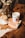 An arrangement of cozy autumn-themed items features a lit candle labeled 'Care Makes' placed on a book, surrounded by dried daisies in a rustic brown bottle and a white dotted teacup. Brown knitted fabric, dried leaves, and a soft fur surface contribute to a warm and inviting atmosphere.