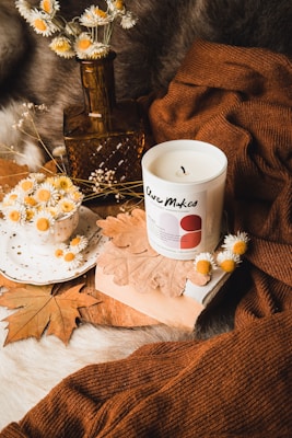 An arrangement of cozy autumn-themed items features a lit candle labeled 'Care Makes' placed on a book, surrounded by dried daisies in a rustic brown bottle and a white dotted teacup. Brown knitted fabric, dried leaves, and a soft fur surface contribute to a warm and inviting atmosphere.