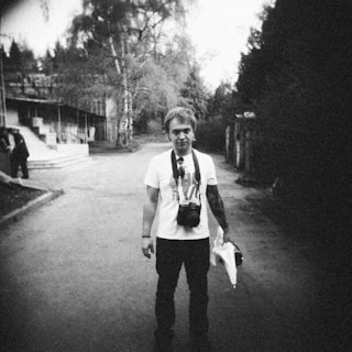 A person stands on a quiet, deserted street. The photograph has a vintage, grainy quality, possibly taken with a film camera. The person is wearing a white t-shirt and dark pants with a camera strapped around their neck. In the background, there are some trees and an old building. The overall setting suggests a peaceful yet slightly melancholic atmosphere.