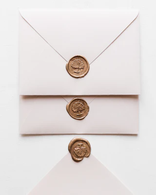 Three pale pink envelopes closed with gold tree-motif wax seals on white cloth