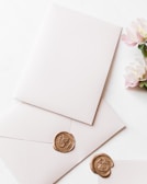 A blank white envelope and matching card are adorned with a gold wax seal featuring an intricate design. Nearby, a small bouquet of light pink flowers adds a delicate touch, contrasting with the soft white background.