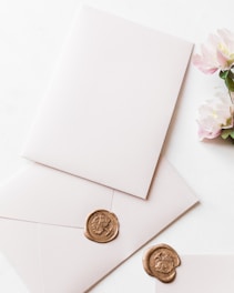 A blank white envelope and matching card are adorned with a gold wax seal featuring an intricate design. Nearby, a small bouquet of light pink flowers adds a delicate touch, contrasting with the soft white background.