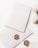 A blank white envelope and matching card are adorned with a gold wax seal featuring an intricate design. Nearby, a small bouquet of light pink flowers adds a delicate touch, contrasting with the soft white background.