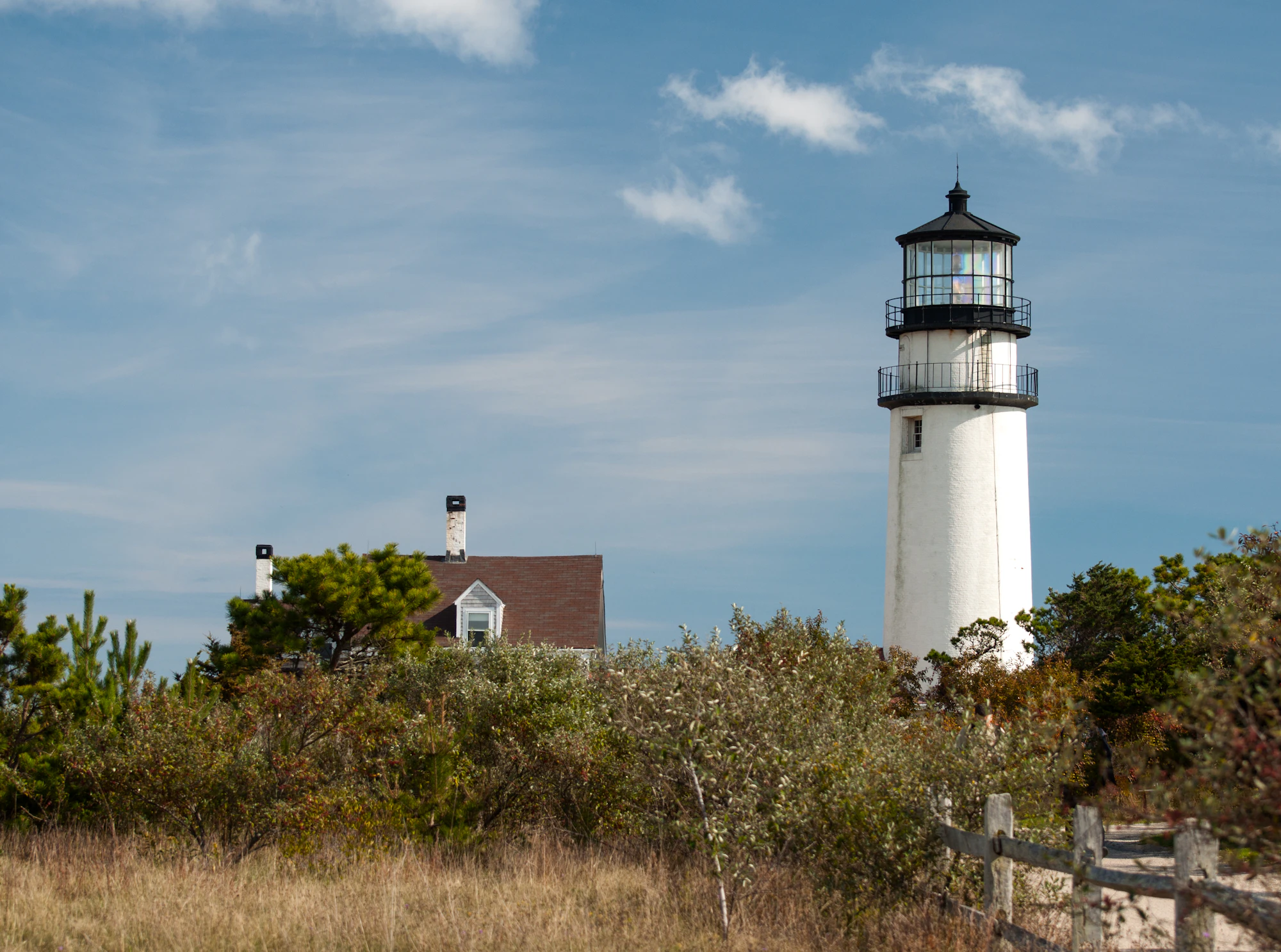 A white lighthouse with black lantern room stands above shrubby dunes under a bright blue sky with scattered white clouds.