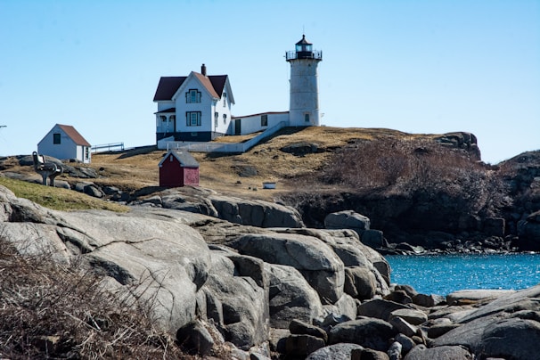 A picturesque coastal scene featuring a white lighthouse with a dark top situated on a rocky outcrop. Nearby, there is a white house with a red roof and additional smaller structures, including a red building. The landscape is rugged with rocks and sparse vegetation. The ocean is visible, displaying a deep blue color under a clear sky.