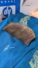 A wooden comb rests on a blue fabric decorated with a yellow feather design. In the background, part of a box with the letters 'dy' in a white and blue color scheme is partially visible.