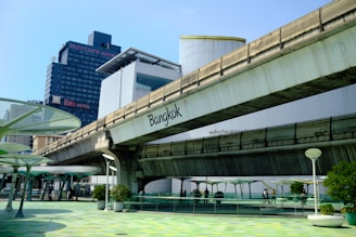 A city scene featuring an elevated concrete bridge with the word 'Bangkok' written on it. In the background, there are tall buildings including a hotel named 'Mercure' and 'Ibis'. The foreground includes a walkway with green and white circular patterns on the ground and modern street lamps. Several plants in pots are placed along the walkway, and some people are visible walking.