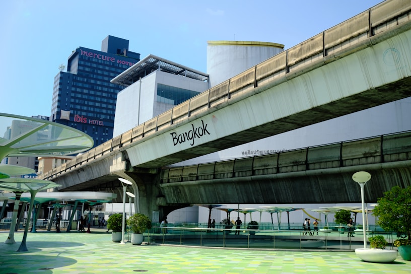 A city scene featuring an elevated concrete bridge with the word 'Bangkok' written on it. In the background, there are tall buildings including a hotel named 'Mercure' and 'Ibis'. The foreground includes a walkway with green and white circular patterns on the ground and modern street lamps. Several plants in pots are placed along the walkway, and some people are visible walking.