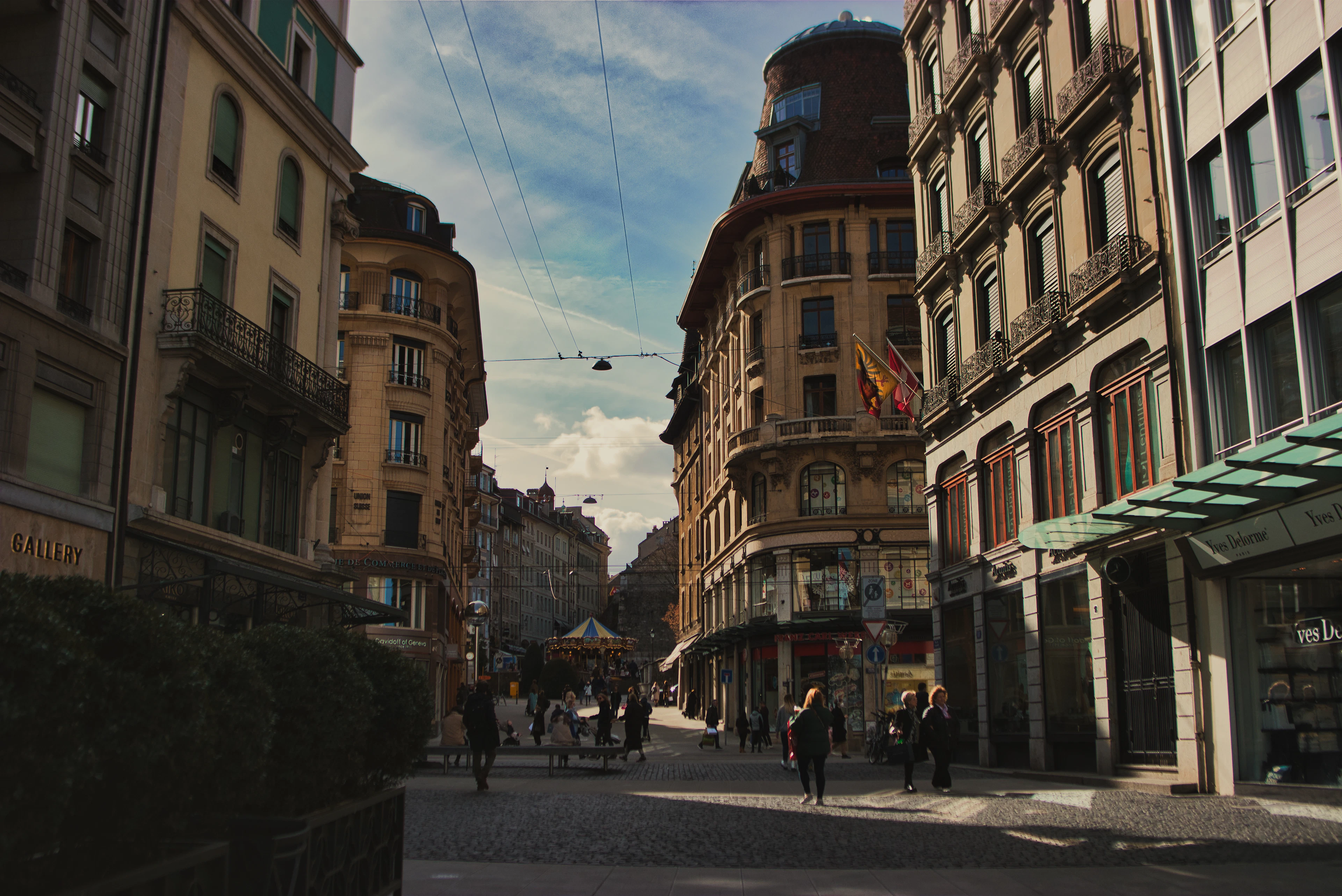 Pedestrians cross a cobbled old-town street in Geneva past a curved corner building and a distant carousel.