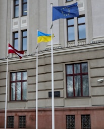 Three flags are displayed on tall flagpoles in front of a beige stone building with large windows and architectural details. The flags include a maroon and white striped flag, a blue and yellow flag, and a blue flag with yellow stars arranged in a circle.
