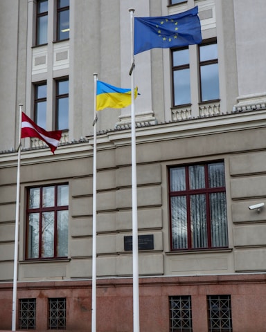 Three flags are displayed on tall flagpoles in front of a beige stone building with large windows and architectural details. The flags include a maroon and white striped flag, a blue and yellow flag, and a blue flag with yellow stars arranged in a circle.