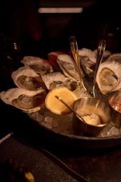 A plate of fresh oysters served on a bed of ice, accompanied by lemon wedges and small metal containers with condiments. The scene is dimly lit, giving it a warm, intimate dining atmosphere.