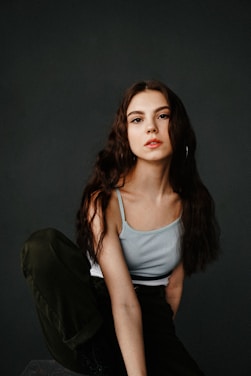 A young woman with long, wavy dark hair poses against a plain dark background. She wears a light blue camisole and dark pants, sitting with one knee raised. Her expression is neutral, with a subtle hint of confidence.