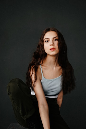 A young woman with long, wavy dark hair poses against a plain dark background. She wears a light blue camisole and dark pants, sitting with one knee raised. Her expression is neutral, with a subtle hint of confidence.