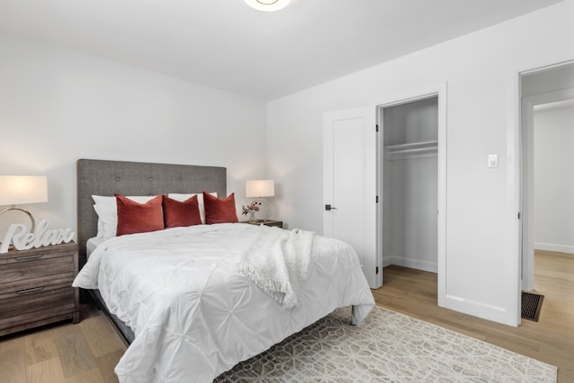 A well-lit bedroom with a neatly made bed featuring white bedding and red accent pillows. The headboard is upholstered in a neutral fabric. On either side of the bed are wooden nightstands with table lamps, and one has the word 'Relax' displayed as a decorative piece. The room includes an open closet with a white door, and the floor is covered with light hardwood.