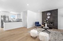 A modern living space featuring an open-plan kitchen and a cozy seating area. The kitchen has white cabinetry and a stainless steel refrigerator, with a black countertop that contrasts with the bright surroundings. The living area includes a sleek black fireplace set against a dark feature wall adorned with decorative circular wall art. A deep blue armchair and fluffy white stools are positioned around a black glass-topped coffee table, which holds a book and a white vase with flowers. The light wood flooring and neutral tones create a clean and inviting atmosphere.
