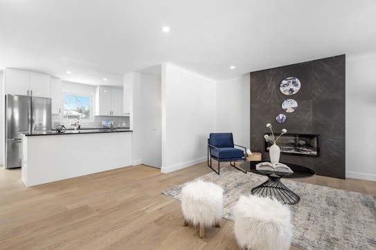 A modern living space featuring an open-plan kitchen and a cozy seating area. The kitchen has white cabinetry and a stainless steel refrigerator, with a black countertop that contrasts with the bright surroundings. The living area includes a sleek black fireplace set against a dark feature wall adorned with decorative circular wall art. A deep blue armchair and fluffy white stools are positioned around a black glass-topped coffee table, which holds a book and a white vase with flowers. The light wood flooring and neutral tones create a clean and inviting atmosphere.