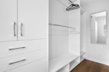 A minimalist white closet with sleek metal handles features open shelving, hanging rods, and a full-length mirror. A few hangers are visible, and there is a window allowing natural light.