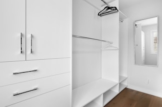 A minimalist white closet with sleek metal handles features open shelving, hanging rods, and a full-length mirror. A few hangers are visible, and there is a window allowing natural light.