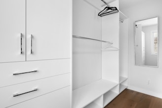 A minimalist white closet with sleek metal handles features open shelving, hanging rods, and a full-length mirror. A few hangers are visible, and there is a window allowing natural light.