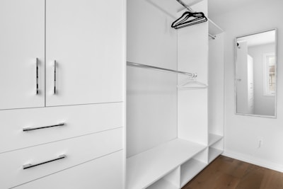 A minimalist white closet with sleek metal handles features open shelving, hanging rods, and a full-length mirror. A few hangers are visible, and there is a window allowing natural light.