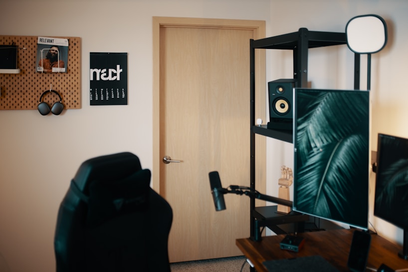 A modern home office setup features a black gaming chair on the left. An acoustic wall panel holds headphones and magazines. A wooden door is centrally placed, and next to it is a black metal shelf with a speaker and some decorative items. Greenery-themed art is displayed on a monitor, with a desk microphone in the foreground.