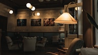 A cozy café interior featuring a dimly lit atmosphere with vintage armchairs and a standing lamp casting a warm glow. The walls are adorned with framed artwork, and modern spherical pendant lights hang from the ceiling. One person can be seen sitting in the background, adding a peaceful, intimate vibe to the space.
