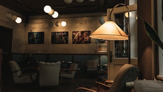A cozy café interior featuring a dimly lit atmosphere with vintage armchairs and a standing lamp casting a warm glow. The walls are adorned with framed artwork, and modern spherical pendant lights hang from the ceiling. One person can be seen sitting in the background, adding a peaceful, intimate vibe to the space.