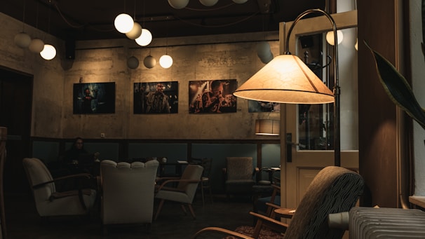 A cozy café interior featuring a dimly lit atmosphere with vintage armchairs and a standing lamp casting a warm glow. The walls are adorned with framed artwork, and modern spherical pendant lights hang from the ceiling. One person can be seen sitting in the background, adding a peaceful, intimate vibe to the space.