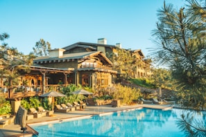 A luxurious resort with wooden architecture surrounded by lush greenery is visible. The main building has a rustic design with multiple levels and balconies. In front of the building, there is a large, inviting swimming pool with lounge chairs and umbrellas arranged neatly beside it. Tall trees and shrubs enhance the natural look of the area, and the sky is clear and blue.