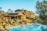 A luxurious resort with wooden architecture surrounded by lush greenery is visible. The main building has a rustic design with multiple levels and balconies. In front of the building, there is a large, inviting swimming pool with lounge chairs and umbrellas arranged neatly beside it. Tall trees and shrubs enhance the natural look of the area, and the sky is clear and blue.