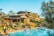 A luxurious resort with wooden architecture surrounded by lush greenery is visible. The main building has a rustic design with multiple levels and balconies. In front of the building, there is a large, inviting swimming pool with lounge chairs and umbrellas arranged neatly beside it. Tall trees and shrubs enhance the natural look of the area, and the sky is clear and blue.