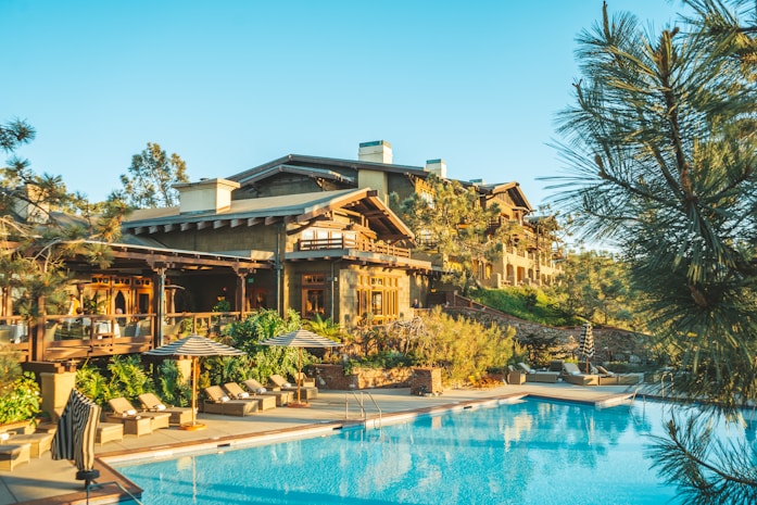 A luxurious resort with wooden architecture surrounded by lush greenery is visible. The main building has a rustic design with multiple levels and balconies. In front of the building, there is a large, inviting swimming pool with lounge chairs and umbrellas arranged neatly beside it. Tall trees and shrubs enhance the natural look of the area, and the sky is clear and blue.
